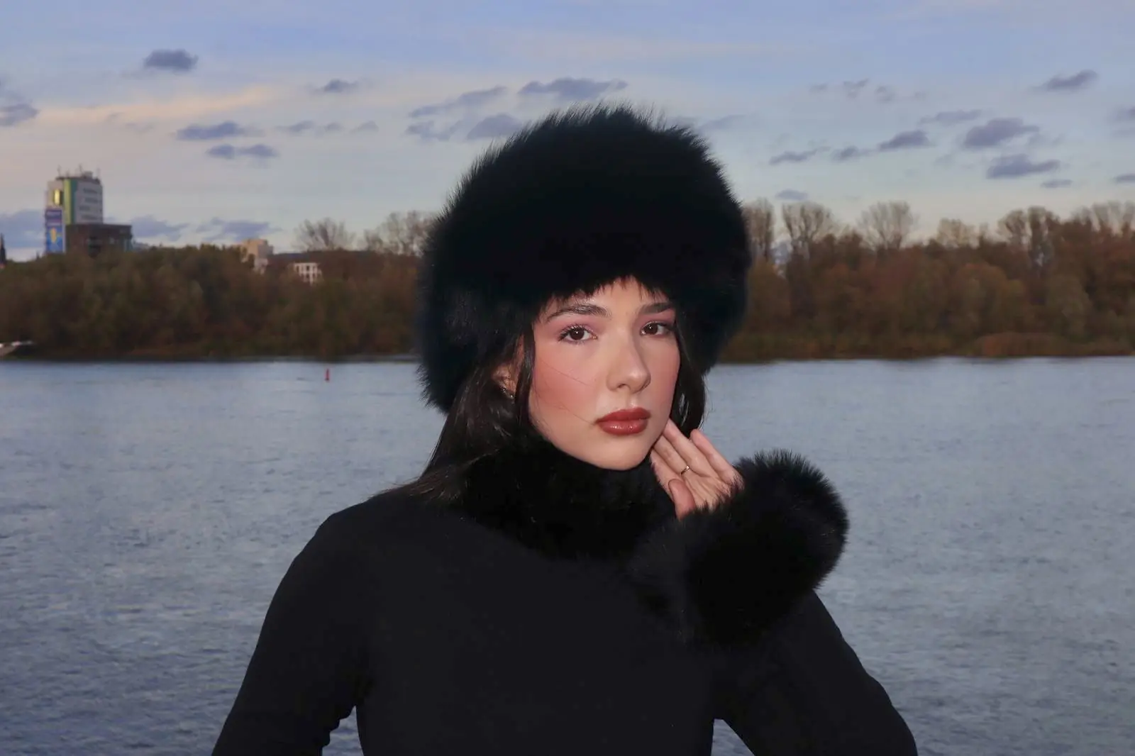 Model in black with a black real fur hat by the river at dusk
